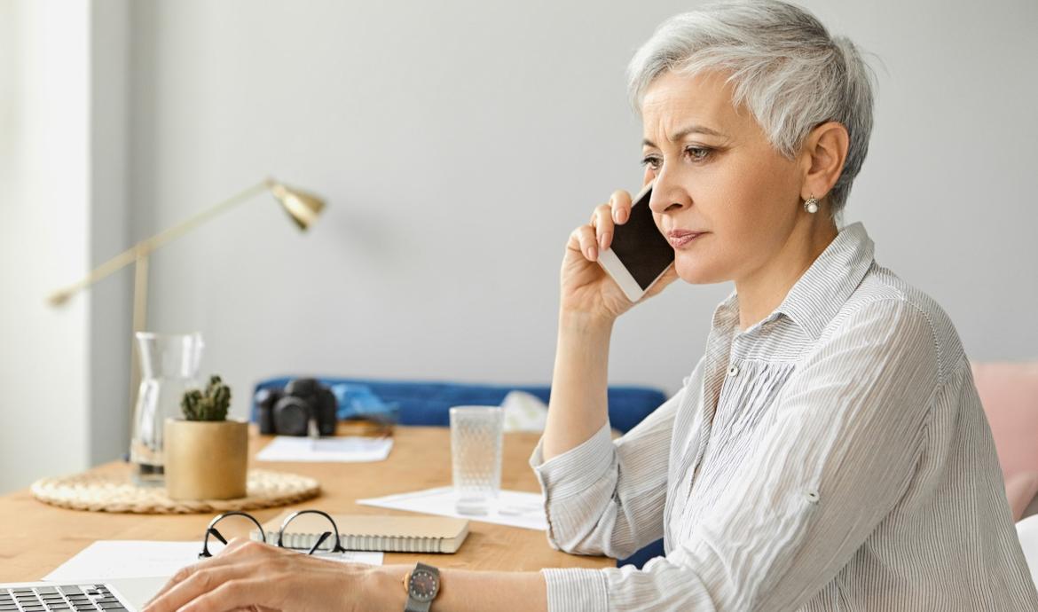 Ältere Frau mit kurzen grauen Haaren telefoniert am Schreibtisch mit ernstem Gesicht