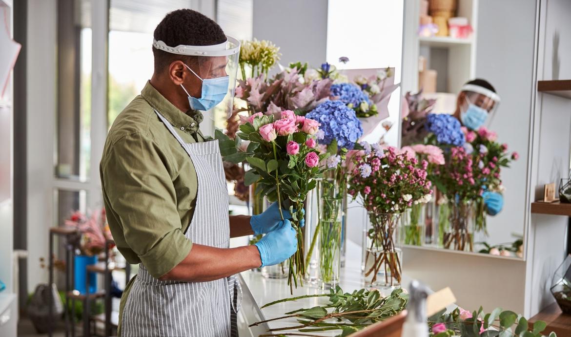 Junger Blumenerkäufer steht mit Maske und Visier und bindet in seinem Laden einem Blumenstrauß
