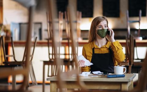 Lokalbesitzerin sitzt im leeren Lokal mit Maske vor Rechnungen und telefoniert deprimiert